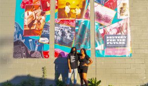 Silent Fox and Olivia House standing in front of a 3 panel photo banner of an art exhibition they worked on with the U of M. 